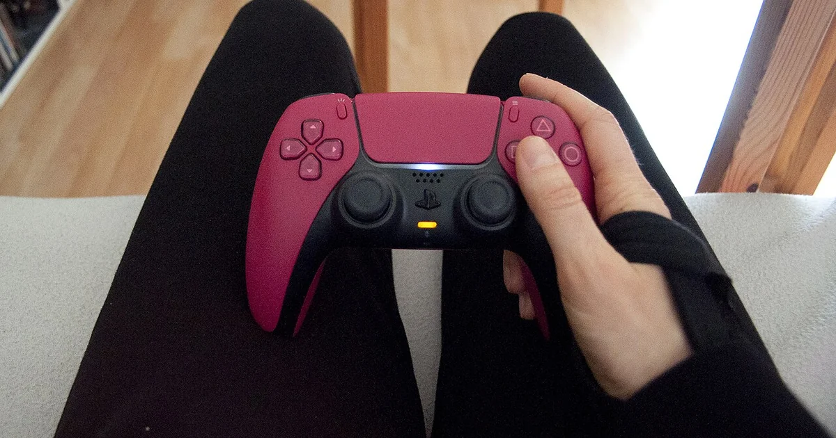 A person sitting on a white couch is holding a pink PlayStation 5 controller in their right hand. They are wearing a black wrist brace on their right wrist. The person has their right index finger on the shoulder button and their right thum